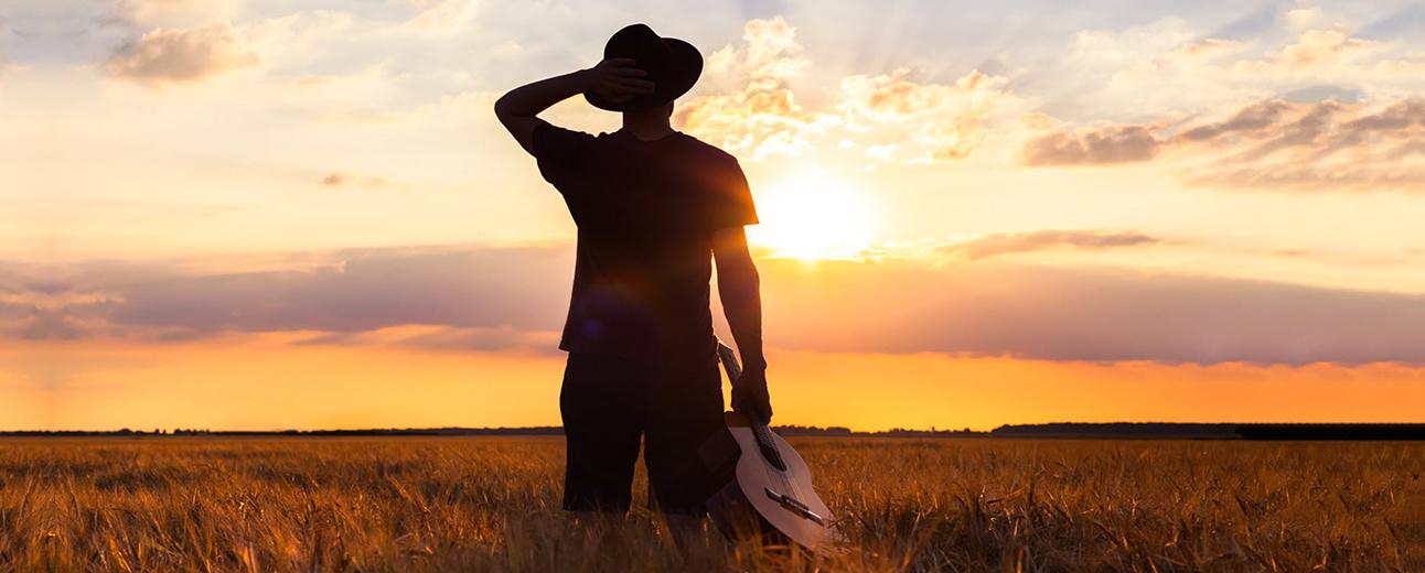 A person standing in a field at sunset, holding a hat and a guitar, looking towards the horizon.